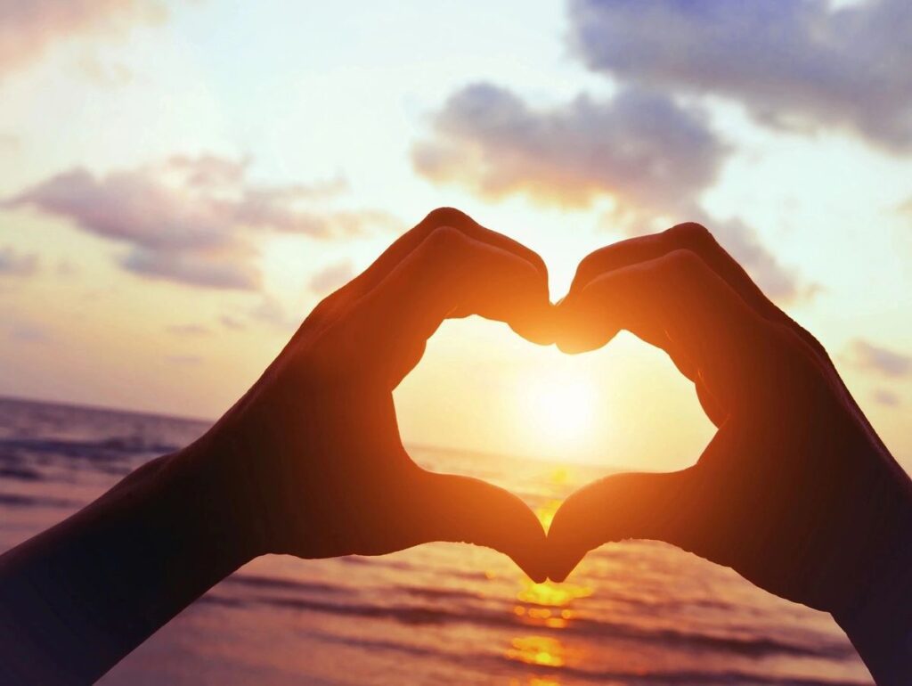 Hands forming a heart shape around the setting sun over the ocean.